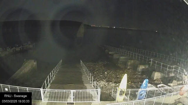 A dark, nighttime scene by the waterfront showing a partially lit pier extending over the water. On the left side, several large rocks are visible along the shore. To the right, two kayaks are stored on the ground, one yellow and one blue. A white railing runs along the edge of the pier, leading to a view of calm, dark water with faint lights in the distance on the opposite shore. The date and time displayed in the bottom left corner indicate March 3, 2025, at 19:00:33.