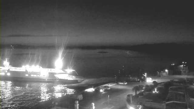 A dark, nighttime scene featuring a large ferry docked at the water's edge, illuminated by bright lights. The water reflects the light from the ferry. In the foreground, several vehicles, including white cars, are parked or moving along a road by the dock. The background shows a calm body of water, with the outline of distant land or islands visible under a dimly lit sky.