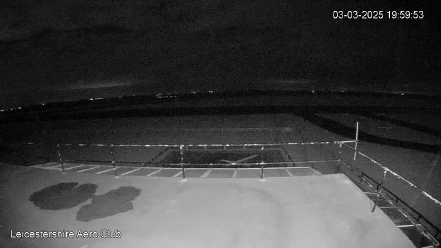 A dimly lit scene captured by a webcam at Leicestershire Aero Club. The image features a flat surface with faint shadows, possibly indicating the presence of objects or markings. In the background, a dark sky is visible, with subtle outlines of distant lights. A railing is seen on the right side, with patterns suggesting the division of spaces. The timestamp in the corner indicates the date and time of the image.