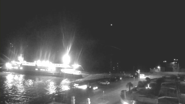 A dimly lit scene at night shows a large ferry at a dock, illuminated by bright lights. The water next to the ferry appears slightly reflective. Various vehicles are parked nearby, with some illuminated by their headlights. In the background, there are distant lights and structures, suggesting a harbor or waterfront setting.