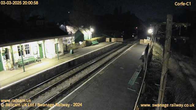 The image shows a dimly lit train station at night. There are two railway tracks running through the center of the image. On the left, there is a stone building with green trim that houses a waiting area. A green bench is visible in front of the building. To the right, there are wooden fence panels and additional benches along the platform. A single light illuminates the area, while the background is mostly dark, containing indistinct shapes of trees or buildings. The image includes a timestamp indicating the date and time at the top.