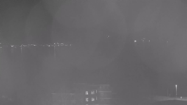 A dimly lit scene captured by a webcam at night. The foreground includes a dark building with some windows illuminated. In the background, there are faint lights reflecting off the water, suggesting a distant shoreline or cityscape. The overall image has a grayish tone, creating a slight foggy or obscured visibility effect.