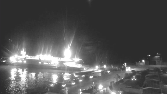 A brightly lit ferry is docked at a harbor at night. The water is calm, reflecting the lights. The dock is lined with vehicles and lampposts, casting soft glows on the surroundings. In the background, some buildings are faintly visible, with additional lights illuminating the scene.