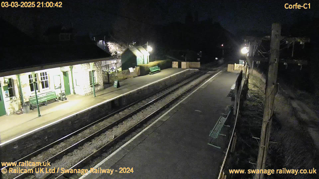 A dimly lit railway station during nighttime, featuring a platform with several benches. On the left side, there is a stone building with windows and a green bench outside. A sign indicates "WAY OUT." The platform stretches alongside two railway tracks that are partly visible. Sparse lighting illuminates the area. In the background, there are trees with some light reflecting off them, suggesting there may be structures or pathways nearby.