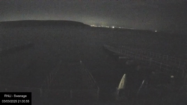 A dark scene of a coastal area at night, with a faint view of distant lights on the horizon. In the foreground, there is a structure that looks like a wooden pier, with some visible seating areas and a partially obscured small boat covered with a tarp. The atmosphere appears calm and quiet, with the water reflecting some of the minimal ambient light.