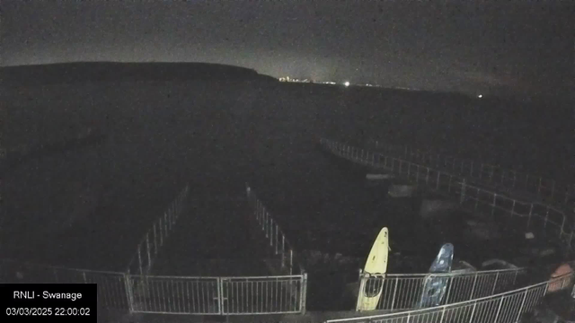 A dimly lit scene at a waterfront captured by a webcam. In the foreground, there are two kayaks, one yellow and one blue, positioned near a low railing. The background features a dark body of water blending into the night sky, with a faint outline of distant lights along the horizon. The image has a dark atmosphere, suggesting it was taken at night. The date and time stamp indicates the image was captured on March 3, 2025, at 10:00 PM.