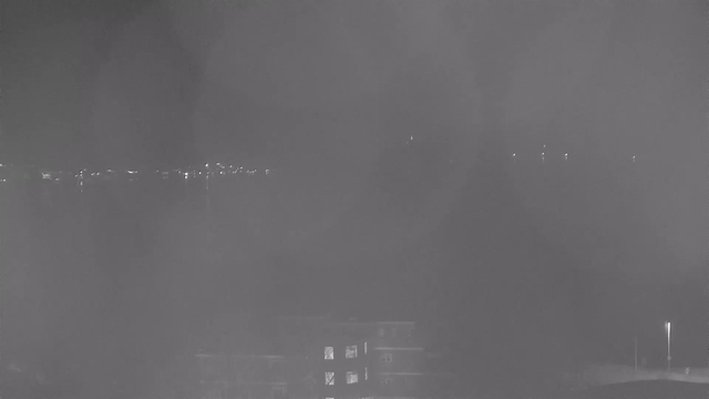 A nighttime webcam image showing a dark scene with faint lights along the horizon, likely from distant buildings or streetlights. In the foreground, a silhouette of a building is visible, with some illuminated windows. The overall image has a misty or foggy appearance, obscuring details further into the scene.