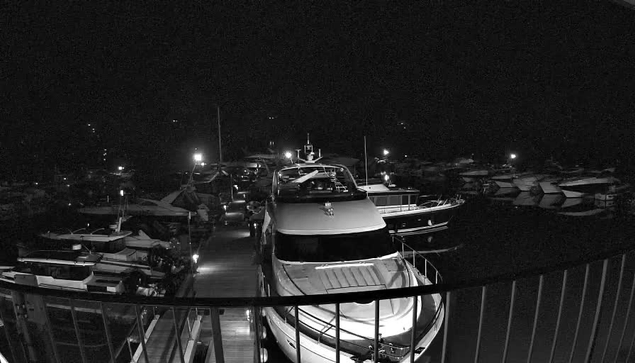 A night scene at a marina, viewed from a balcony. Multiple boats are docked along the water, with some illuminated by distant lights. The foreground features a large white yacht, with its top and deck visible, while the background includes several other boats partially obscured by darkness. The overall atmosphere is calm and quiet, with a hint of activity suggested by the positioning of the boats.