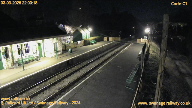 A dimly lit railway station at night, featuring a stone building with green trim and windows. A green bench is visible on the platform, and there are two track lines with gravel in between. On one side, there is a sign indicating "WAY OUT" and a fenced-off area with another green bench. In the background, there are trees and a light source illuminating part of the scene. The platform is empty, suggesting a quiet atmosphere.