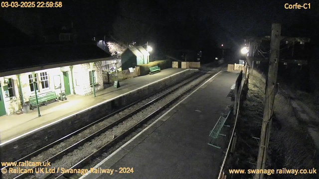 A dimly lit railway station at night is visible. The platform includes several green benches and a sign indicating "WAY OUT." On one side, there is a stone building with large windows and a canopy overhead. A background of dark trees can be seen beyond the station. There are two sets of railroad tracks leading away from the platform, with a wooden fence lined along the edge. A light fixture illuminates part of the area while the night sky is dark.