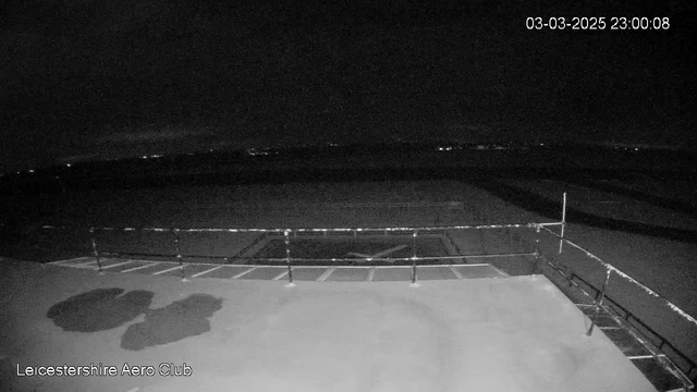 A nighttime image captured from a webcam at Leicestershire Aero Club, showing a dark scene with very few lights visible in the distance. The foreground features a moored railing with a partially visible paved area that has markings indicative of an aerodrome, including a large “X” shape on the surface. The ambiance is quiet and subdued, with the date and time displayed in the upper right corner.