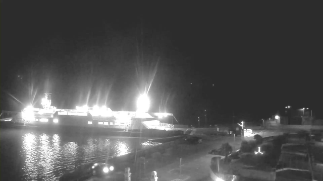 A brightly lit ferry at a dock, with its ramp extending toward the shore. The surrounding area is dark, illuminated by streetlights and the ferry's lights reflecting on the water. The scene appears quiet, with only a few vehicles and structures visible along the dock.