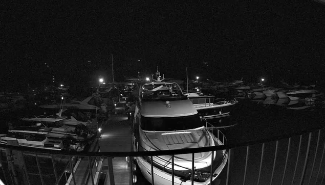 A nighttime image of a marina filled with various boats. A large yacht is prominently in the foreground, with a dock leading away from it. The scene is illuminated by artificial lights from the boats and docks, creating reflections on the water. The background has numerous smaller vessels, and the overall atmosphere is calm and quiet, typical of a marina at night.