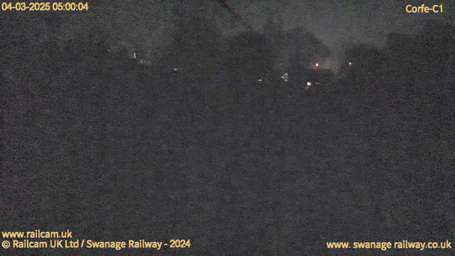 A dimly lit scene captured by a webcam, showing a dark landscape with vague shapes and two small sources of red light in the background. The time stamp in the top left corner reads "04-03-2025 05:00:04." The image appears grainy and lacks detail, suggesting a nighttime setting with very low visibility.