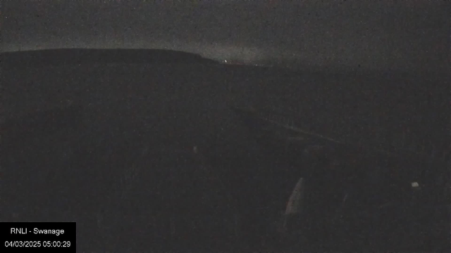 A dark scene captured by a webcam, showing a shadowy landscape with hills in the background. The lighting is very dim, with faint outlines of the terrain visible. In the far distance, there are small scattered lights, likely from buildings or boats. The overall atmosphere is nighttime, creating a quiet and still ambiance.