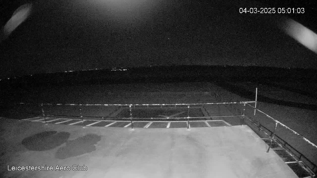 A dark scene captured from a webcam at Leicestershire Aero Club, displaying an empty airstrip. The ground is mostly flat and appears to be wet, likely from moisture or rain. In the background, faint lights can be seen, suggesting distant structures or vehicles. A railing is visible along the edge of the platform, leading towards the airstrip. The image is timestamped, indicating it was taken just after dawn.