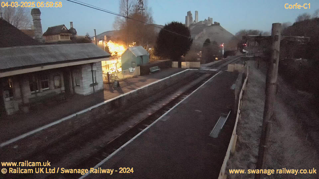 A dimly lit scene of a railway station in the early morning. The foreground shows the railway tracks running through the image with a wooden fence on the right. On the left, there is a station building with a sloped roof and several windows. A bench is located nearby. In the background, a small blue structure is visible, illuminated by warm lights. Above, there are power lines and a mountain with a castle or fortress structure rising in the distance. The overall atmosphere is quiet and tranquil, with a hint of dawn light in the sky.