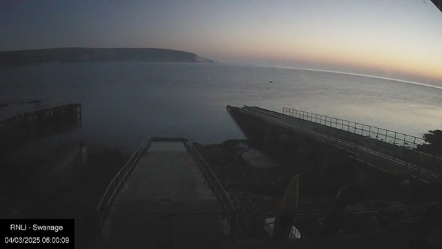 A tranquil seaside scene at dawn, with a calm ocean reflecting soft orange and blue hues in the sky. A jetty extends out into the water from the left, and to the right, a rocky shoreline is visible. The area is empty, creating a serene and quiet atmosphere.