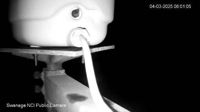 A close-up view of a white cylindrical object mounted on a wooden platform. A white cable extends from the object, and there are metal bolts visible beneath the platform. The background is dark, suggesting it is early morning or nighttime. A timestamp in the corner indicates the image was captured at 6:01 AM on March 4, 2025.