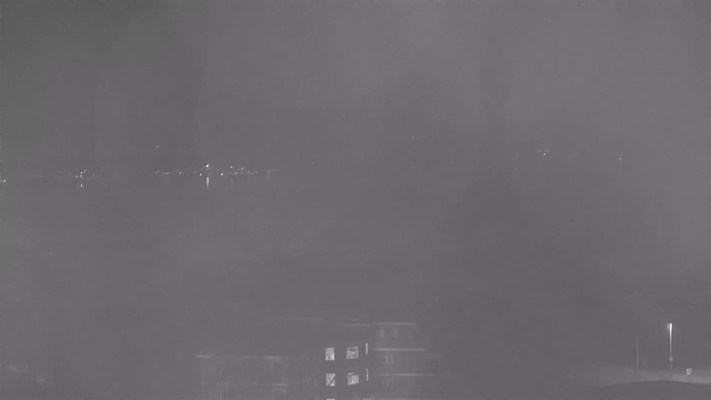 A dark, monochromatic webcam image shows a hazy view of a waterfront at night. There are faint, distant lights on the horizon, suggesting the presence of buildings or a city skyline. In the foreground, the outlines of structures are visible, with one building displaying a few illuminated windows. The overall scene appears dim and atmospheric, with a sense of obscurity and distance.