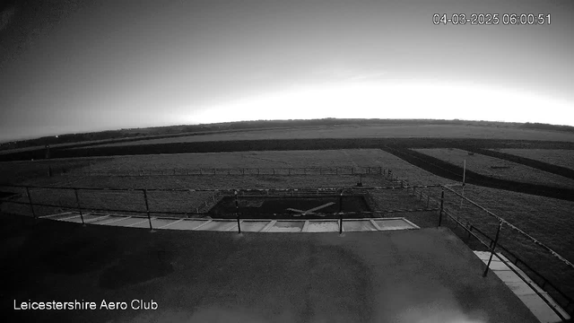 A black and white webcam image shows a runway area at the Leicestershire Aero Club. The foreground features a flat surface with a railing along the edge. The runway stretches out toward the horizon, surrounded by open fields. The sky appears clear and gradually lightening, indicating early morning. A timestamp in the corner reads 04 March 2025, 06:00:51.