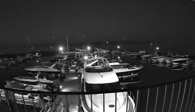 A nighttime view of a marina filled with various boats moored closely together. The scene is illuminated by multiple lights reflecting off the water, creating a serene and tranquil atmosphere. A wooden dock runs through the marina, with a few figures visible, suggesting activity. The sky is dark, enhancing the contrast between the illuminated areas and shadows of the boats.