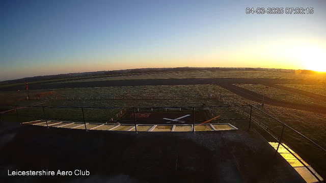 A sunrise scene over a grassy airfield. In the foreground, there is a terrace railing with a view of the runway, which has a small aircraft outlined on it. In the background, a clear blue sky transitions into warm yellow and orange hues from the rising sun. The landscape is mostly flat, with distant hills visible under the morning light. The airfield is bordered by a fence, and there are no clouds in the sky. The image is timestamped at 7:00 AM on the 4th of March, 2025.