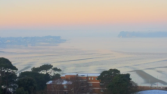 A serene view of a calm body of water at sunrise, with soft pastel colors in the sky transitioning from pink to light orange. In the foreground, a brown building with a flat roof is partially visible, surrounded by green trees. The water reflects the gentle hues of the sky and features subtle ripples. In the distance, there are hints of land and possibly structures along the shoreline, shrouded in a light mist. The overall atmosphere is tranquil and peaceful.