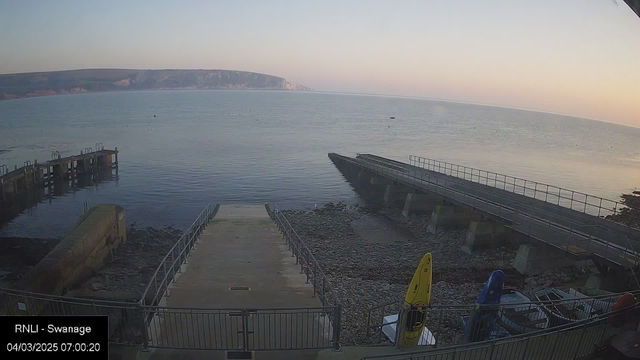 A calm sea view with gentle waves reflects the soft light of dawn. In the foreground, a boat ramp leads down to the water, flanked by a rocky shoreline. To the right, a small pier extends into the water, with a few boats moored nearby. Yellow and blue kayaks are parked on the shore, and the coastline is visible in the distance, with cliffs softly illuminated by the early morning light. The sky transitions from pale blue to warmer hues near the horizon.