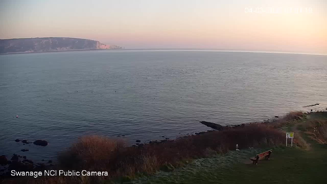 A coastal scene at dawn, featuring calm waters and a distant cliff. The sky transitions from soft pink to pale blue as the sun rises. In the foreground, grass and a few bushes frame the scene, alongside a wooden bench. A sign and a few scattered rocks are also visible near the shore. The atmosphere is peaceful and serene, with no people present.