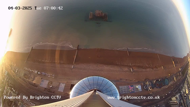 A high-angle view captured by a webcam shows a sandy beach with a calm sea, reflecting morning light. In the water, remnants of a pier can be seen, partially submerged. The shoreline has a few wooden structures and reflects the absence of people. On the left, there are faint outlines of buildings and possibly an amusement area, while the edge of the photo captures the curvature of a glass structure at the top of the frame. The time and date are displayed in the upper corner.