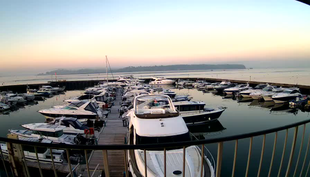 A scenic view of a marina at dawn, featuring numerous boats docked in still water. The boats vary in size and type, with some covered and others visible in detail. The sky has soft hues of orange and pink as the sun rises. In the background, land is visible with trees and possibly a structure. The overall atmosphere is calm and serene.