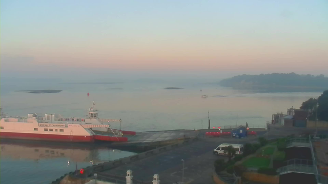 A calm seascape at dawn features a ferry docked at a harbor. The ferry has a white body with a red bottom and signage indicating "Give Way to Chain Ferry." The water appears still and is reflecting soft pastel colors of pink and blue from the sky. In the distance, there are small boats and a distant shoreline with trees outlined against the horizon. The nearby harbor area has a few vehicles, a blue building, and landscaped greens, with gentle waves breaking against the shore.