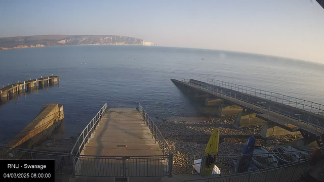 A serene coastal scene showing a wide view of calm water reflecting the sky. In the foreground, there are two piers extending into the water, one curved and the other straight, with several boats visible. The shoreline is rocky and features a ramp leading down to the water. On the right side, there are colored kayaks propped against the railing. The background includes cliffs rising above the water, and the sky is clear, indicating a bright morning.