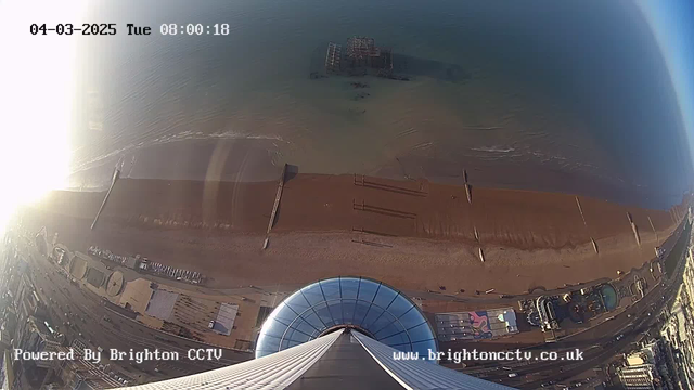 A high-angle view of a sandy beach and the ocean taken from a webcam. In the foreground, the lens shows part of a circular structure, possibly an observation deck or building rooftop. Below are sandy sections of the beach, with some wooden planks and a pier extending into the water. In the distance, there is a submerged structure in the ocean. The left side of the image is bright, likely due to sunlight, while the right side shows a clearer view of the beach and water. The time and date display is in the upper left corner, indicating it is early morning on a Tuesday.