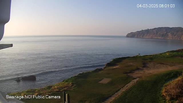 A calm coastal scene featuring a vast, smooth sea reflecting the light of the early morning sky. On the left side, there is a slight land protrusion into the water with a dark rock partially submerged. The shoreline in the foreground is grassy with a few patches of dry land visible, and a stone pathway leading toward the water. In the background, a distant cliff rises above the horizon, partially obscured by soft morning light. The timestamp on the top right corner indicates the date and time of the image.