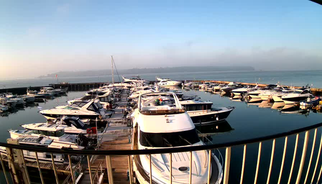 A view of a marina with numerous boats docked. The scene features a calm water surface reflecting the boats and a light blue sky. There are several larger motorboats and smaller vessels arranged along the docks. Some boats have visible sails and covered areas, while others appear sleek and modern. The background shows a hazy shoreline and hills, suggesting a peaceful morning atmosphere. A railing is visible in the foreground, framing the scene.