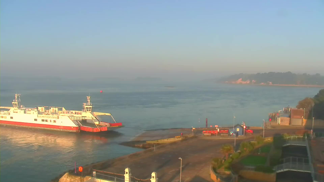 A ferry with a red and white hull is docked at a port beside calm waters. The background features a clear blue sky with some haze and distant land visible. To the right, there is a small blue building and a pathway leading to the waterfront. Various red barriers and a few trees are present near the dock, with sunlight reflecting off the water's surface.