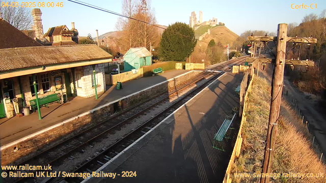 A well-lit outdoor scene shows a railway station platform with wooden benches painted green. The station building, made of stone with a sloped roof, sits to the left. In the background, a castle is visible on a hill, surrounded by trees. The sky is clear and blue, indicating a sunny day. There is a wooden fence with an opening marked "WAY OUT" on the right side of the image. Shadows cast by the benches create patterns on the platform surface.