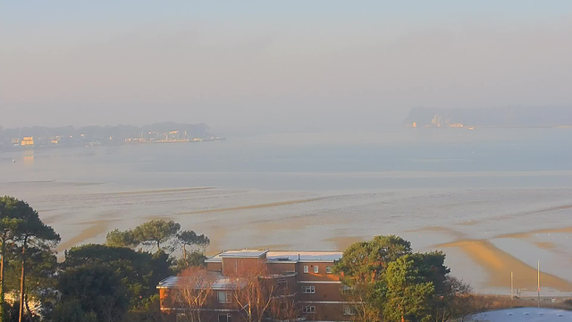 A foggy view over a calm bay in the early morning. In the foreground, there are trees and a low-rise building with several windows. The water stretches out in soft, muted colors of blue and gold, with gentle ripples reflecting the faint light. In the distance, there are small boats and a shoreline dotted with distant buildings against a hazy backdrop of hills. The atmosphere is serene and tranquil, with a subtle mist hanging over the scene.