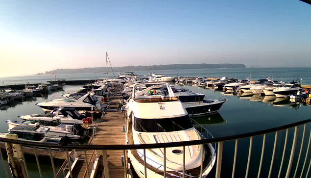 A marina scene featuring numerous boats docked in calm waters, with a wooden walkway visible in the foreground. The sky is clear and bright, and a distant shoreline can be seen in the background. The boats vary in size and design, with some displaying colorful accents.