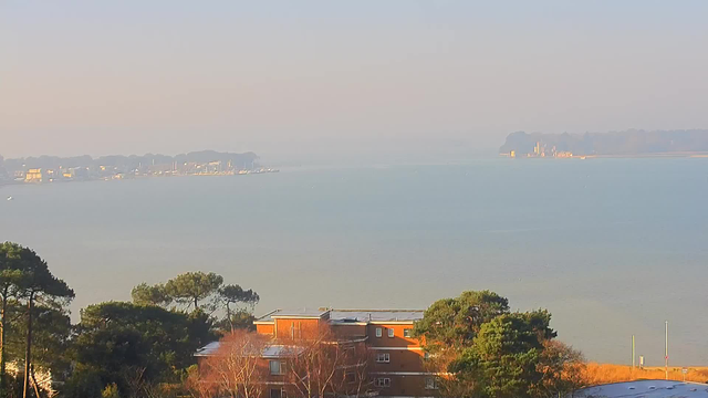 A panoramic view of a calm body of water under a hazy sky. In the foreground, there are trees and low buildings with muted colors. The background features a shoreline dotted with boats and a distant landmass, possibly an island, which is partly obscured by fog. The overall scene conveys a tranquil, serene atmosphere.