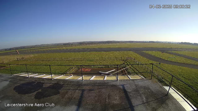 A clear blue sky is overhead with no clouds. In the foreground, a runway is visible, accompanied by a small white airplane positioned on a grassy area. There is a fence perimeter around the grass, and additional grassland extends in the background. The scene is bright and sunny, indicating a pleasant day.