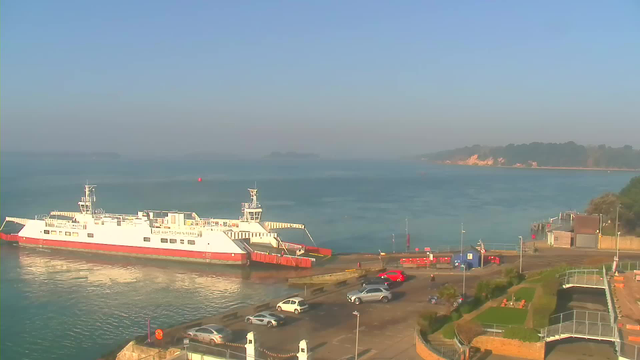 A white ferry is docked at a pier, with its ramp lowered to the terminal. The water is calm, reflecting the blue sky above, while hazy land is visible in the distance. Several vehicles are parked nearby, including a red car and a silver SUV. There are some green areas with seating and a small building on the right side of the image.