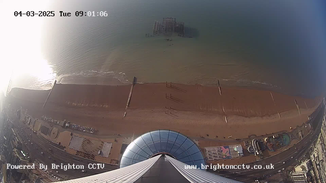 Aerial view of a sandy beach with the ocean visible. In the foreground, the blue dome of a structure is shown, along with lines leading down to the beach area. The beach has a few people scattered along it, and a pier can be seen in the water. There are various structures and a road present on the left side of the image, indicating a coastal area. The date and time stamp is displayed at the top left corner. The light from the sun creates a reflection on the water, enhancing the brightness of the scene.