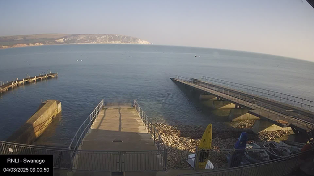 A calm seaside view featuring a concrete dock extending into the water. The surface of the sea is smooth and reflects the light of a clear sky. In the background, distant white cliffs rise above the shoreline, partially shrouded in morning mist. A few boats can be seen floating in the water, and the foreground includes a small sandy area with pebbles. A yellow kayak is resting on the shore, along with several other vessels secured near the dock.