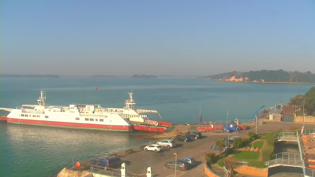 A ferry is docked at a harbor with a red and white color scheme. The water is calm and a light blue, reflecting the clear sky above. In the background, green hills are visible along the shoreline. Various parked cars are situated along the harbor, and some buildings and greenery are present near the edge of the water. Several boats can be seen further out in the water, and a few buoys are also visible. The environment is sunny and bright.