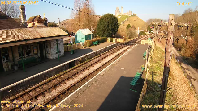 A sunny view of a railway station with several empty benches along the platform. To the left, there is a stone building with a sloped roof, and behind it, a green shed. In the background, a hill features a castle ruin. The railway tracks extend toward the horizon, with gravel filling the space between them. A wooden pole with electrical wires stands on the right side, and a pathway is visible on the far right leading into a grassy area. The sky is clear and blue.