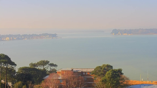 A serene view of a calm body of water under a hazy sky. In the foreground, there are trees and a brown building with multiple windows. The water reflects a soft blue hue, while the distant shoreline is lined with boats and a small marina. Faint silhouettes of buildings can be seen on the horizon, along with a noticeable structure on the right. The scene is tranquil and bathed in gentle light, suggesting early morning or late afternoon.