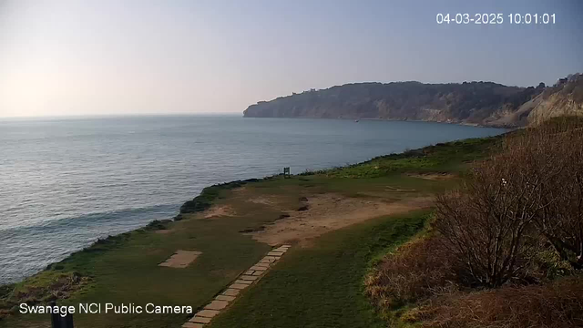 A coastal view captures calm water gently lapping at the shore. In the distance, a cliff rises, partially obscured by a light mist. The foreground features a grassy area with a sparse path made of stones leading towards the water. There are some sparse bushes to the right of the image. The sky is bright and clear, indicating a sunny day. In the lower left corner, the text "Swanage NCI Public Camera" is displayed. The image timestamp reads "04-03-2025 10:01:01."